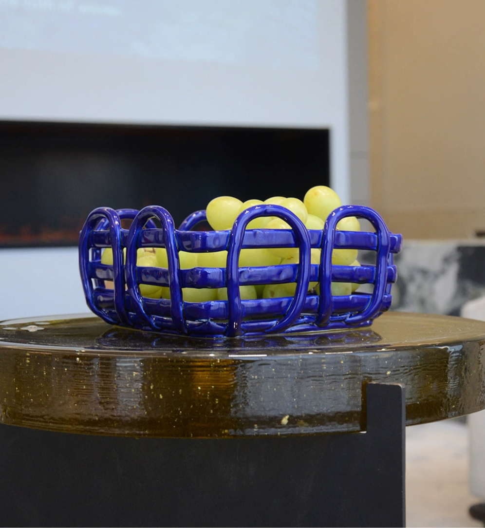 Blue ceramic fruit basket with woven design; vibrant blue color; holds green grapes; sits on a dark, speckled surface.