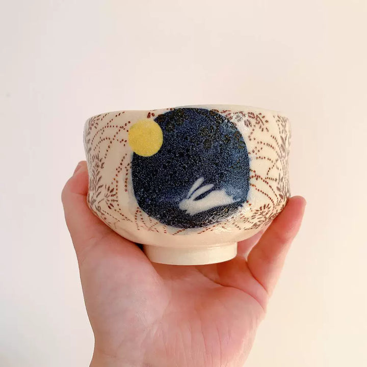 Hand holding a Japanese matcha bowl with a dark blue circle design featuring a white rabbit.