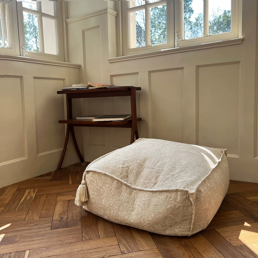 Beige embroidered ottoman pouf with tassel on wood floor, home decor accent.