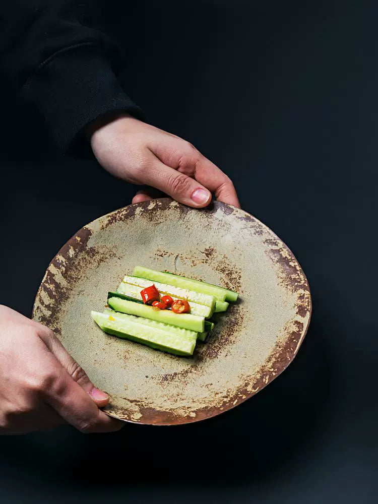 Hands holding a rustic, earth-toned ceramic plate with sliced cucumbers and red chili garnish.
