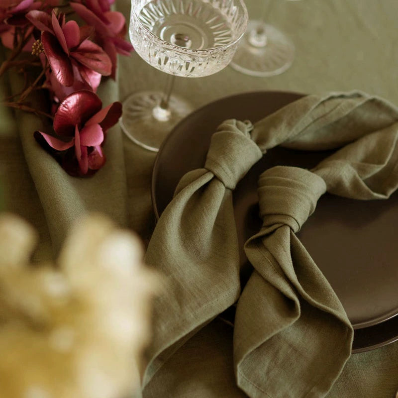 Elegant Linen Table Napkins, knotted in olive green, set on dark plates with floral accents.
