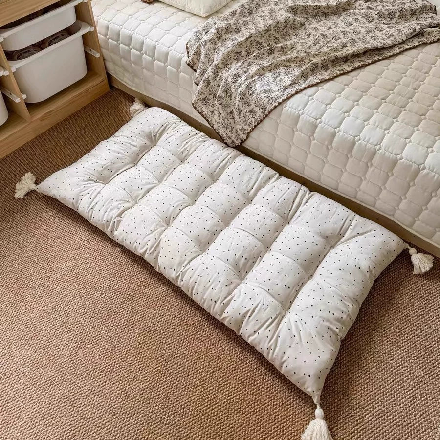 Beige dotted floor cushion with tassels on carpet, beside a bed.
