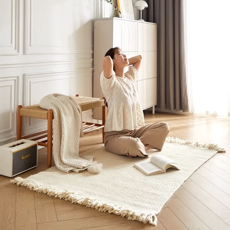 Off-white braided wool rug with fringe, styled with a bench, speaker, and book in a bright room.