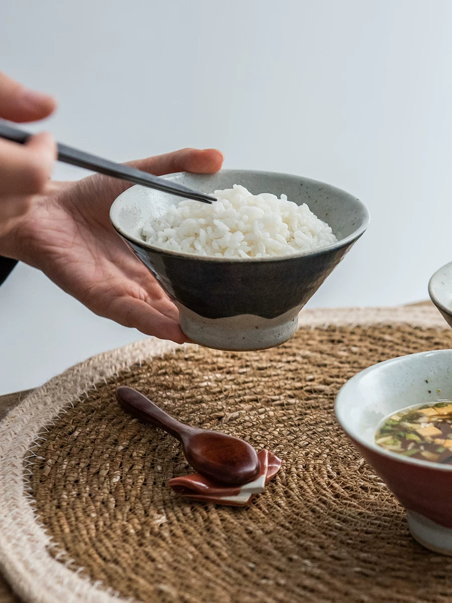 Hands holding a bowl of white rice with chopsticks over a woven placemat.