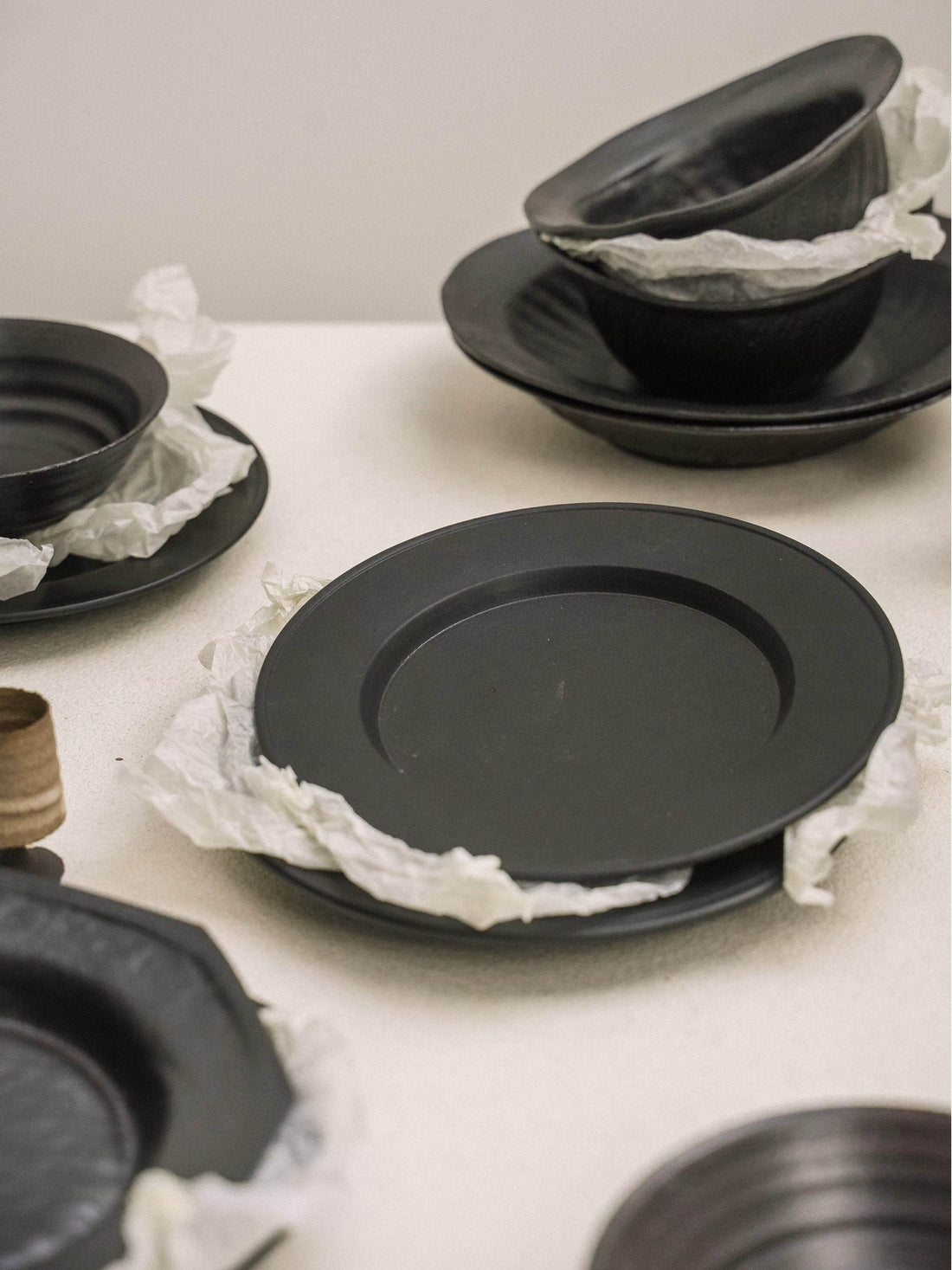 Stack of minimalist matte black ceramic plates and bowls wrapped in protective white tissue paper.