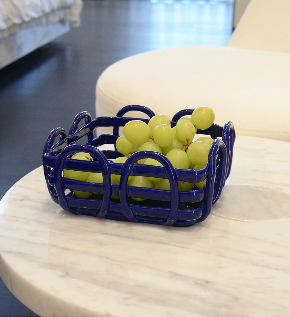 Blue ceramic fruit basket with woven design; filled with green grapes; on a white marble surface.