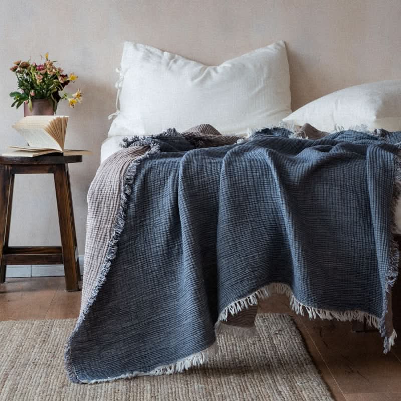Blue and tan Breathable Cotton Throw Blanket with fringe draped on a bed.