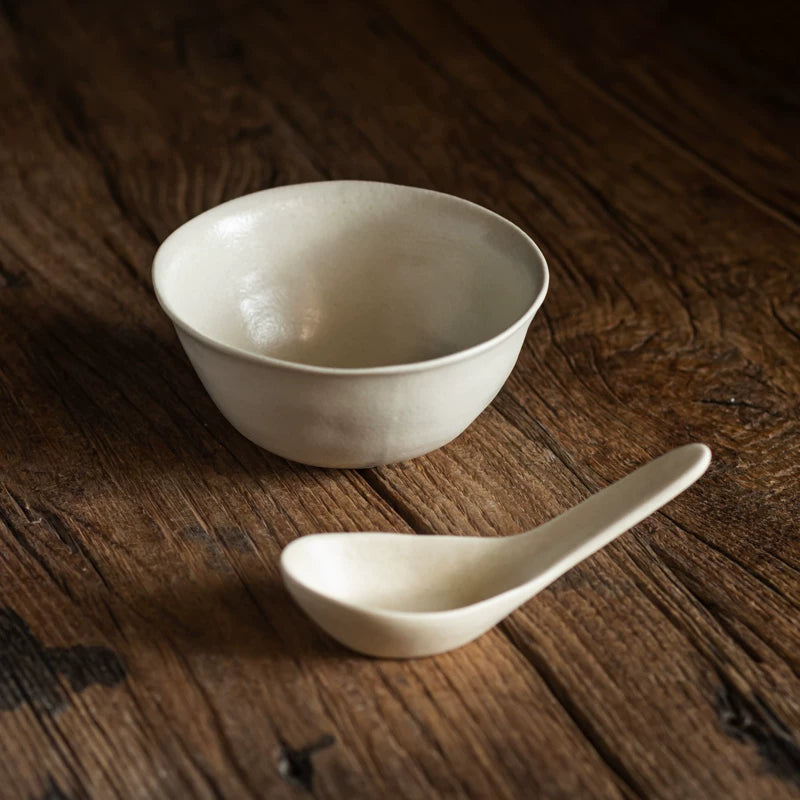 Single creamy white ceramic soup bowl and matching spoon resting on a dark rustic wooden surface.