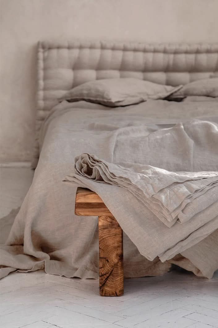 Natural linen bed sheets in beige draped over a wooden bench with a tufted headboard.