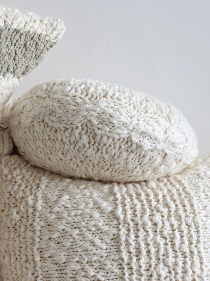 Stacked cream knitted throw pillows, cable knit texture, decorative.