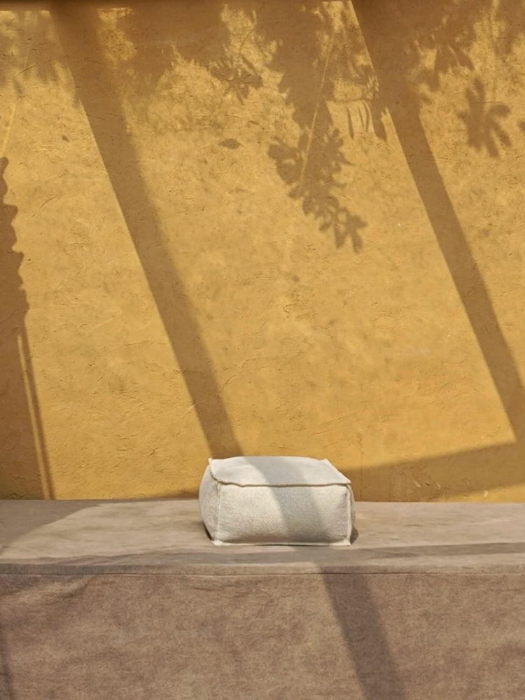 Light beige knitted pouf ottoman, square shape, on a light brown surface, against a mustard yellow wall with shadows.