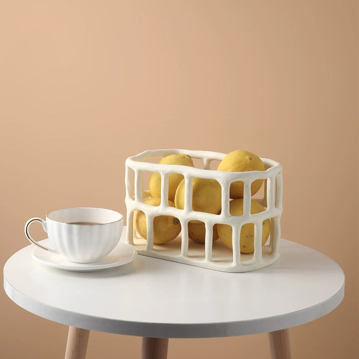 Off-white ceramic fruit basket with a textured, openwork design.  Lemons are inside the basket.  A decorative piece for a kitchen or dining area.