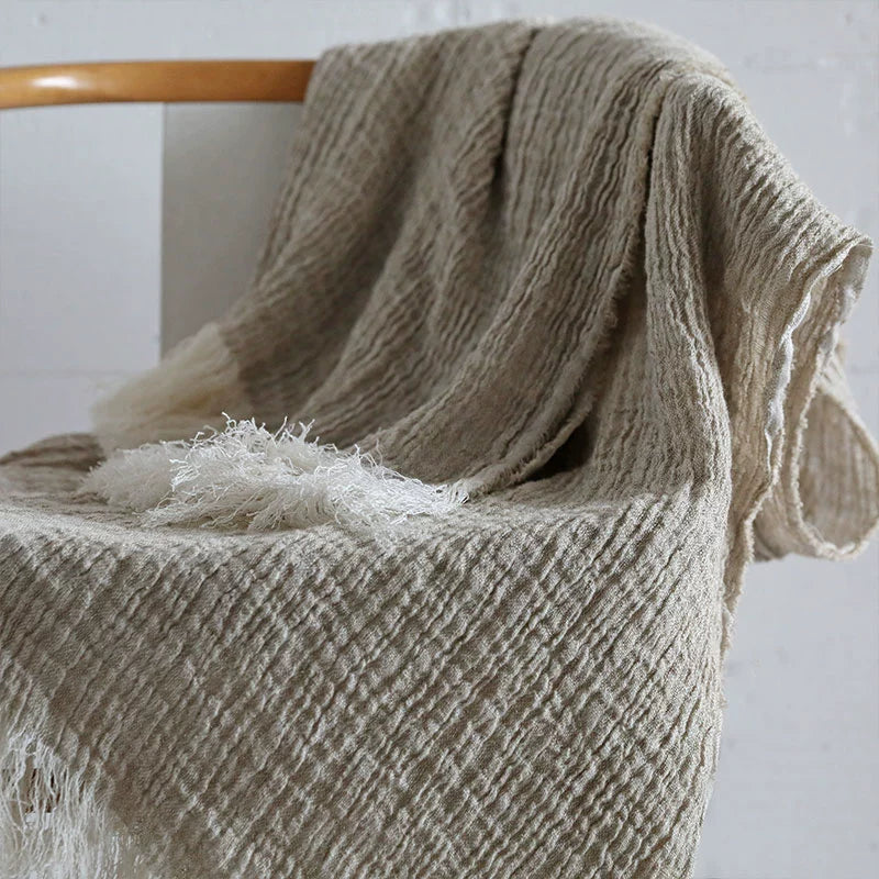 Natural linen throw blanket with white fringes, draped on a chair. Cozy rustic decor.