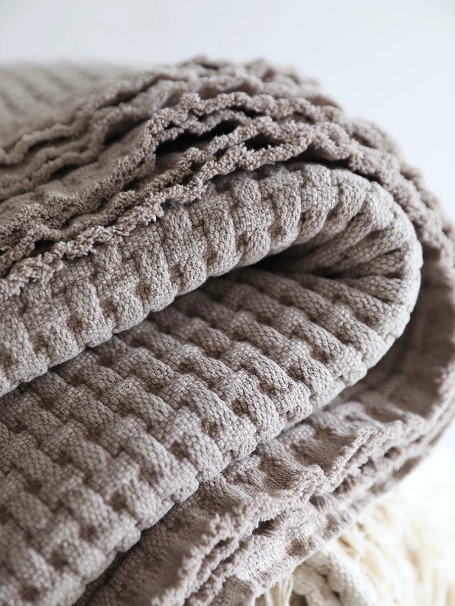 Folded beige woven cotton throw blanket, textured with a repeating pattern.  Fringed edges.