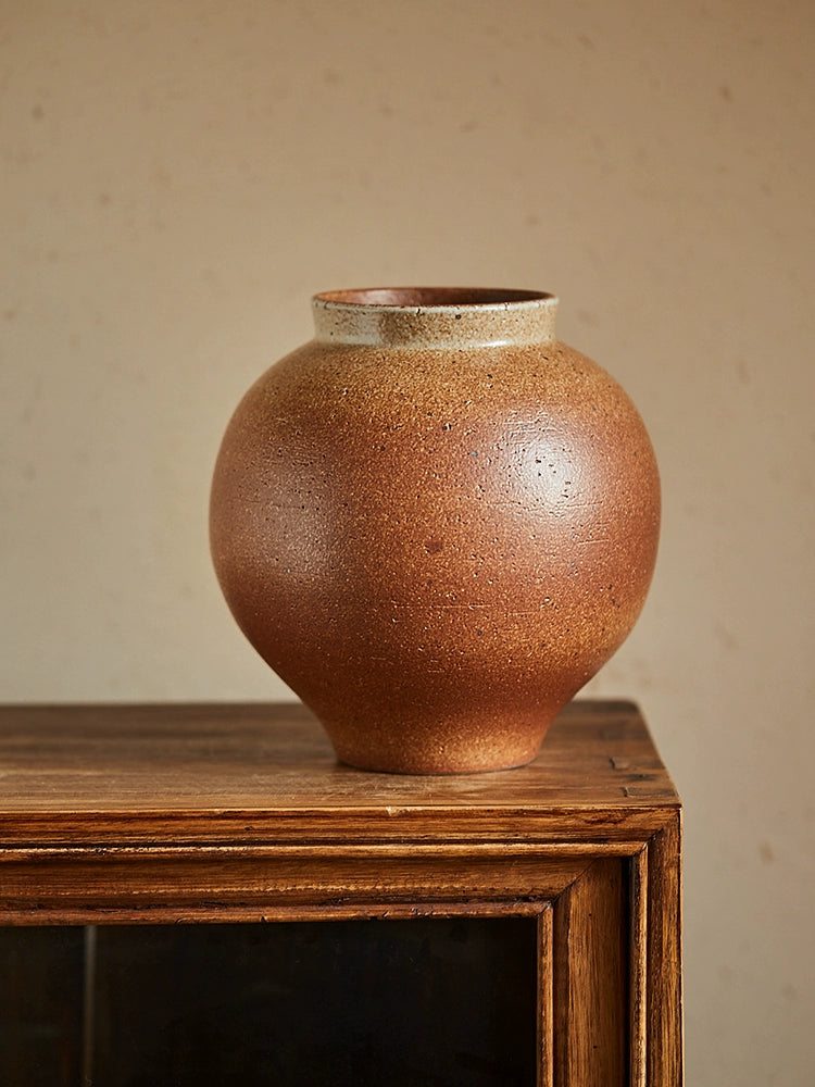 Rustic brown handmade pottery vase, with a speckled, textured surface and a light beige rim.  On a dark wooden cabinet.
