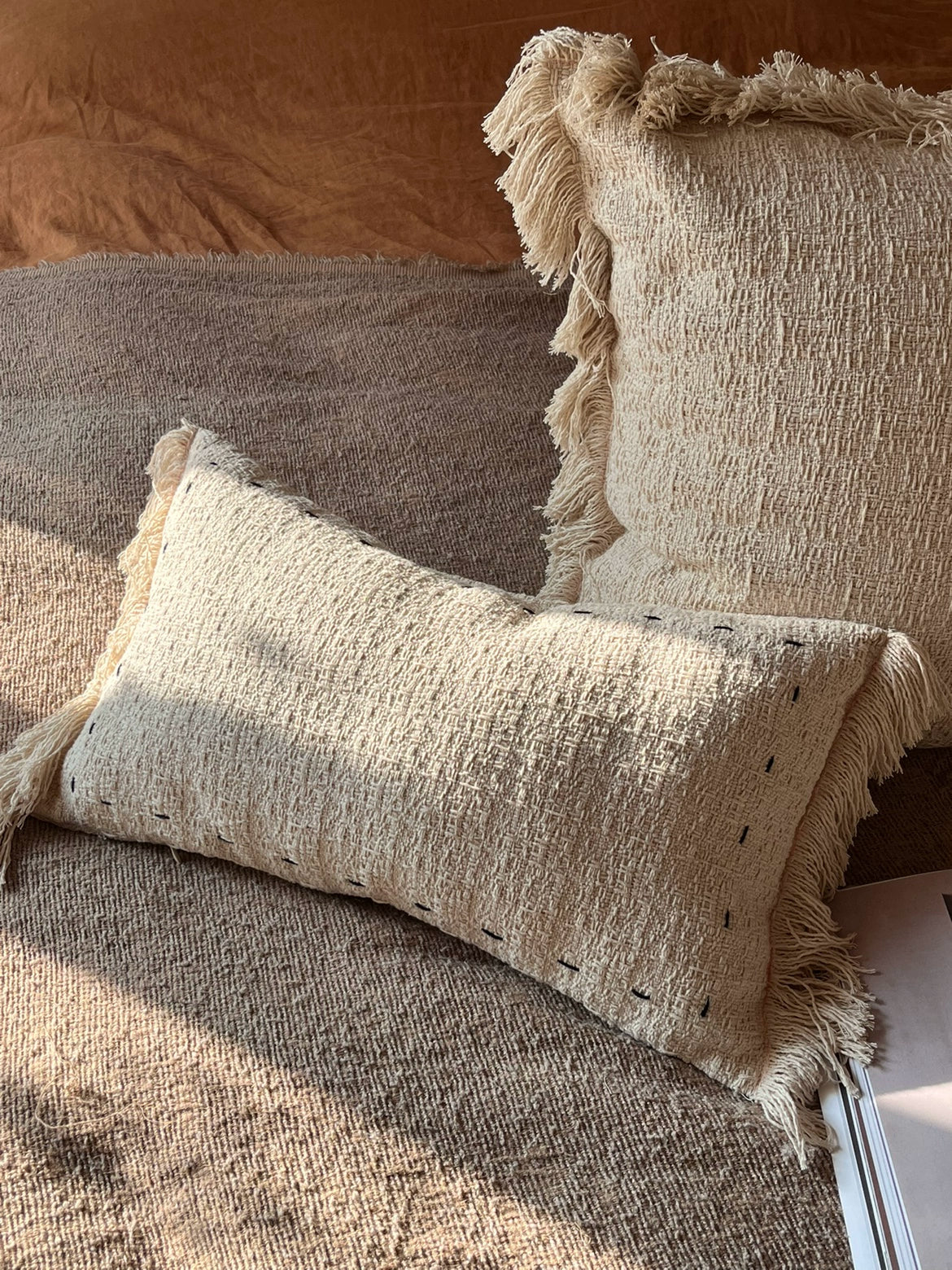 Beige woven throw pillows with fringes, set of 2, on a textured surface.