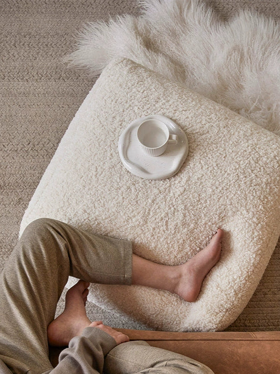 Fluffy cream-colored bean bag chair cushion, square shape, on a beige floor with fur rug.  Person's feet resting on the cushion.  Cozy, soft, plush.