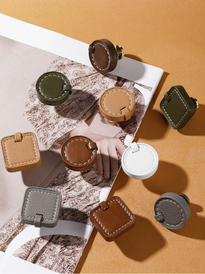 Assorted round and square leather drawer pulls and cabinet knobs in brown, beige, white, dark gray, and green.  Stitched design, various styles.