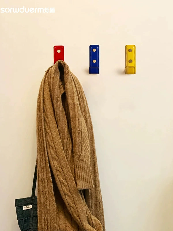 Red, blue, and yellow leather wall hooks hang a brown cardigan. Dopamine decor.