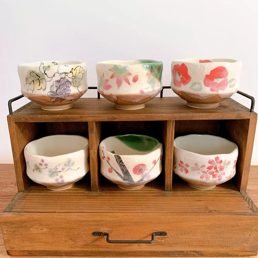 Six floral Japanese matcha bowls displayed in a rustic wooden organizer shelf.