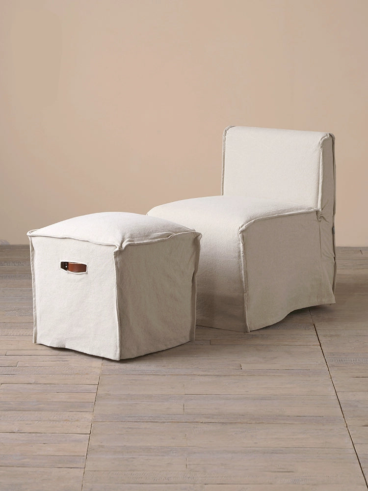 Beige linen square footstool ottoman with leather handle on wooden floor.