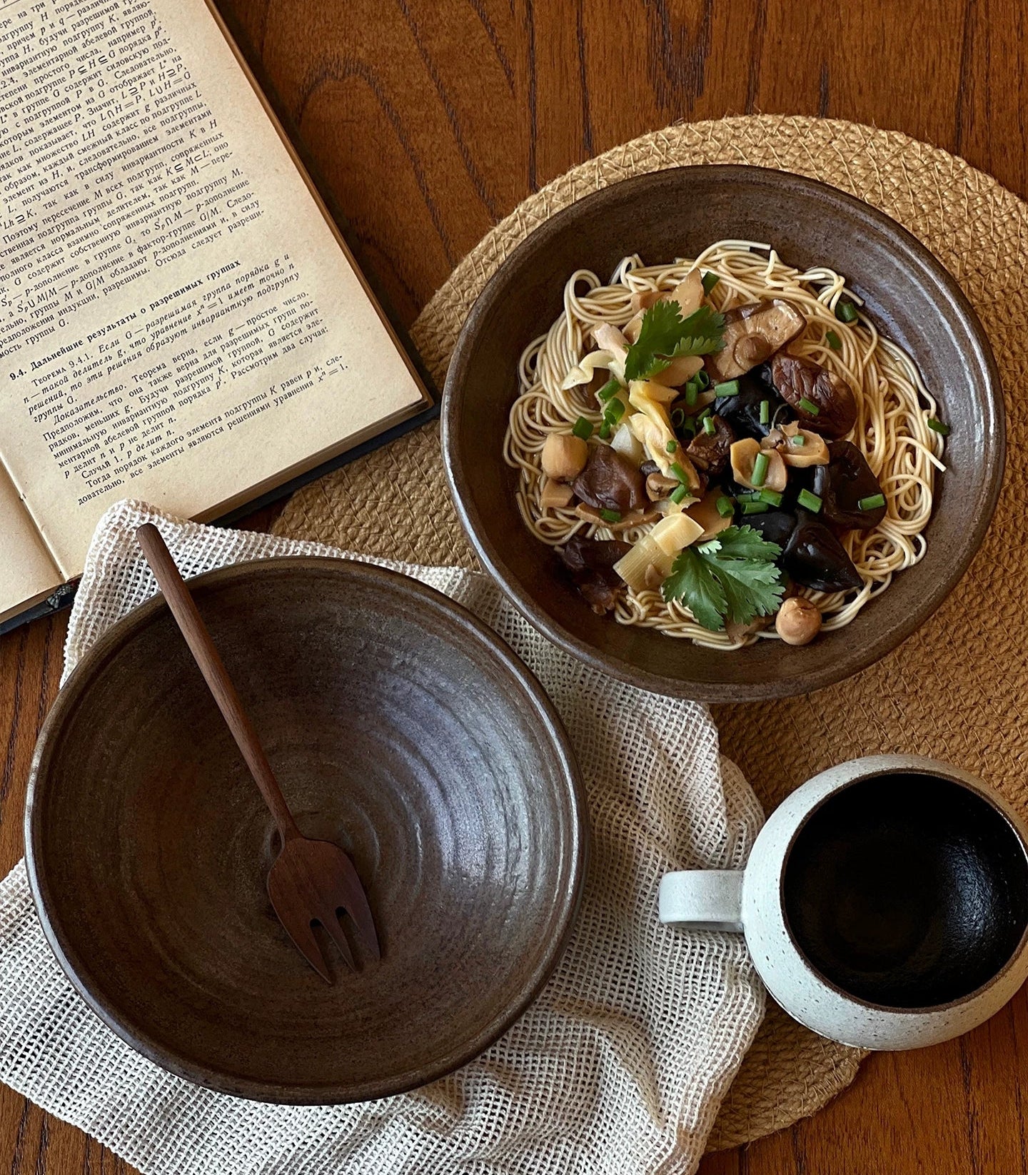 Top-down view of a dark brown ceramic bowl with noodles, next to an empty bowl, a fork, and a white mug.