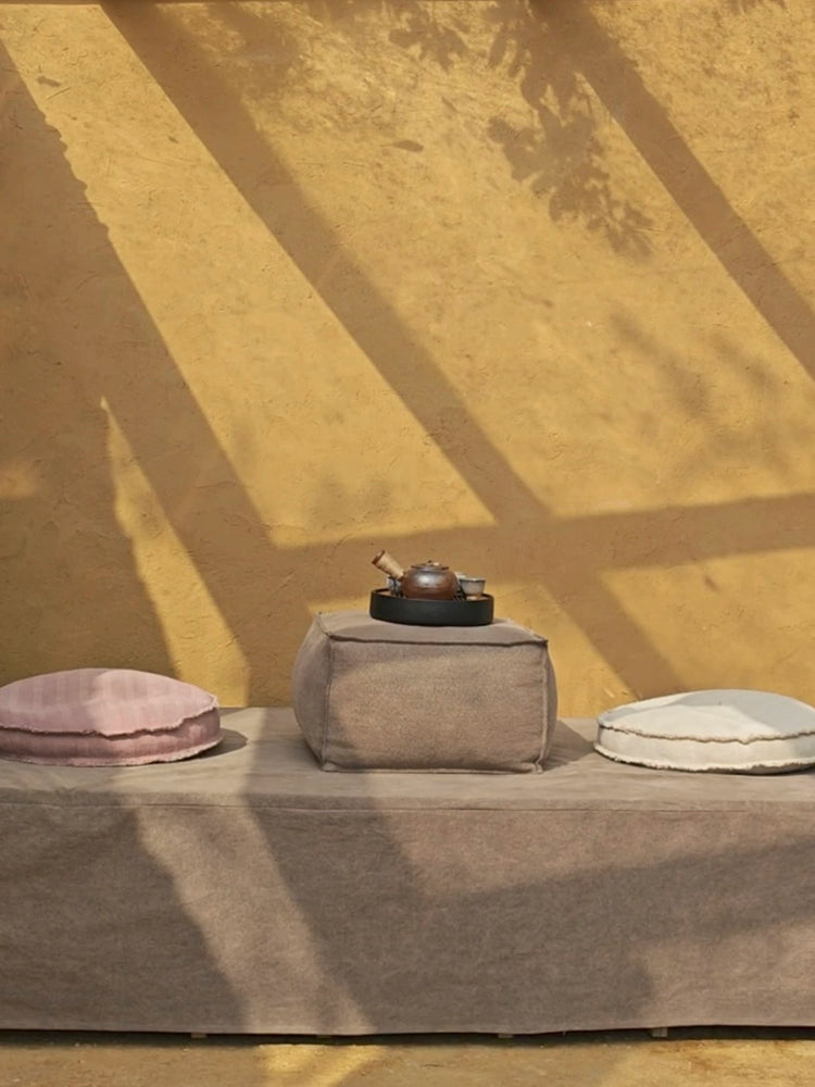 Three square knitted poufs, light taupe, dusty rose, and cream, on a light taupe bench.  Outdoor setting with warm sunlight and shadows.