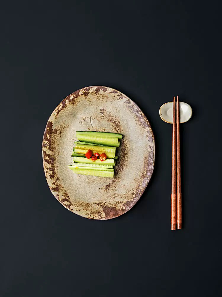 Rustic ceramic plate with sliced cucumbers and chili sauce, next to copper-toned chopsticks on a stand.