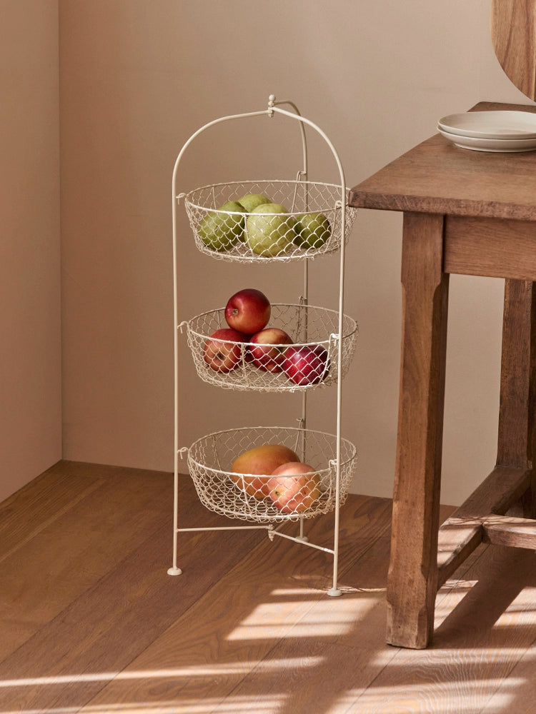 Cream-colored 3-tier wire fruit basket stand with metal wire baskets holding apples, peaches, and pears.  Rustic style kitchen organizer.