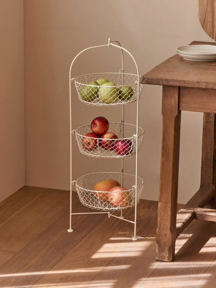 Cream-colored 3-tier wire fruit basket stand with metal wire baskets holding apples, peaches, and pears.  Rustic style kitchen organizer.