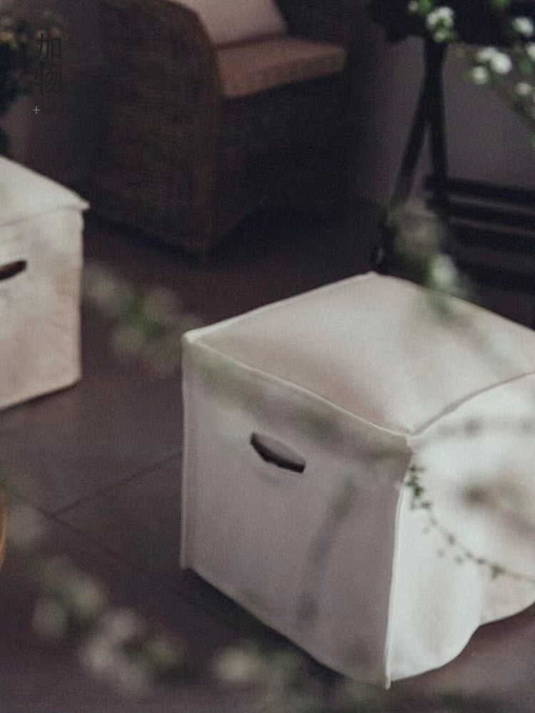 Beige linen square footstool ottoman in a minimalist living room setting.