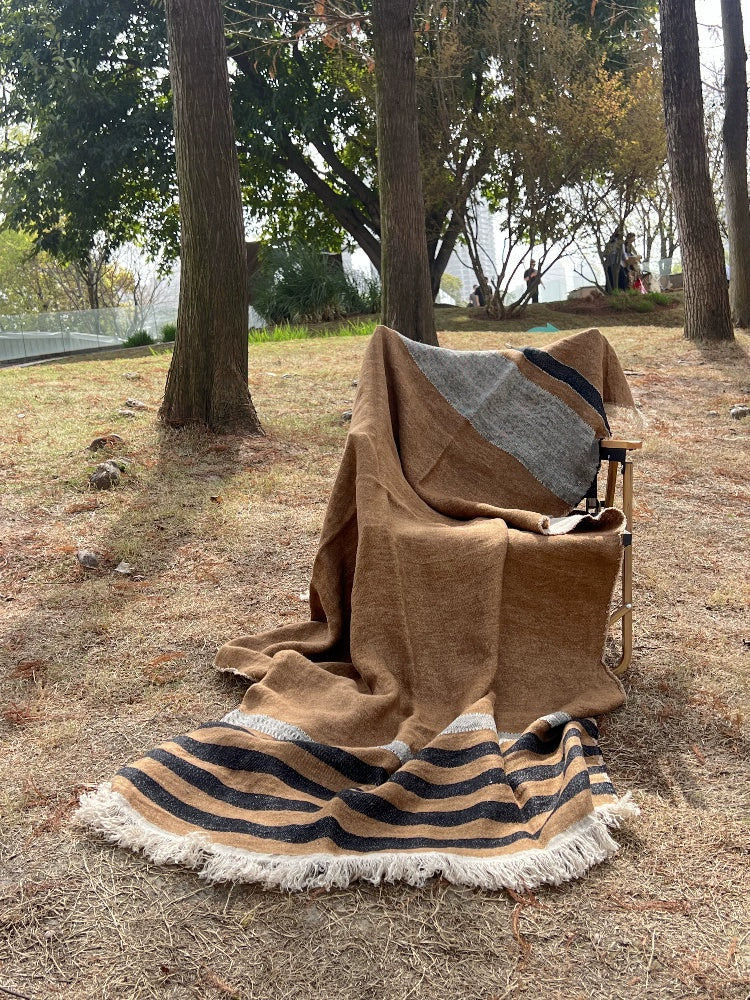 Cozy striped linen throw blanket in brown, black, and gray with fringe, displayed outdoors.