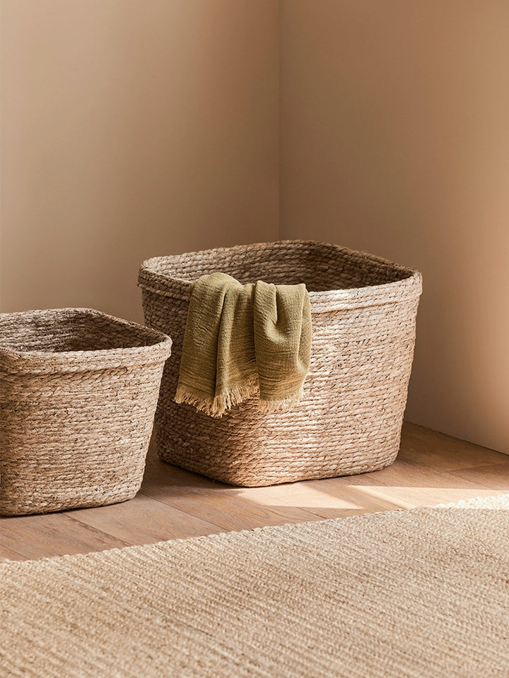 Two natural rattan storage baskets, round and square, on a light beige floor.  Beige-toned woven baskets with a textured surface.  A sage green throw is inside the square basket.