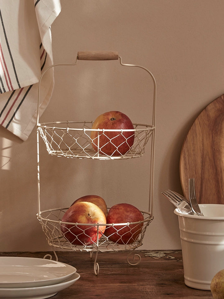 Two-tiered wire fruit basket stand, light beige wire, wooden handle, filled with peaches and apples. Rustic kitchen decor.