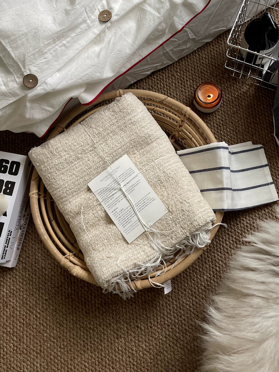 Beige knitted throw blanket, boho style, in a woven bamboo basket.  Fringed edges, soft texture.