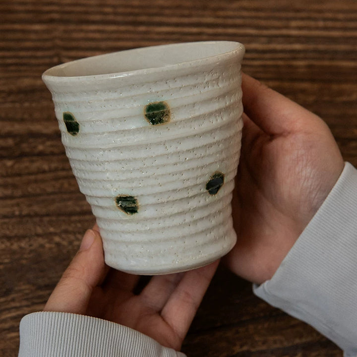Hand holding a textured, white ceramic Yunomi tea cup with dark green polka dots, shot close up.