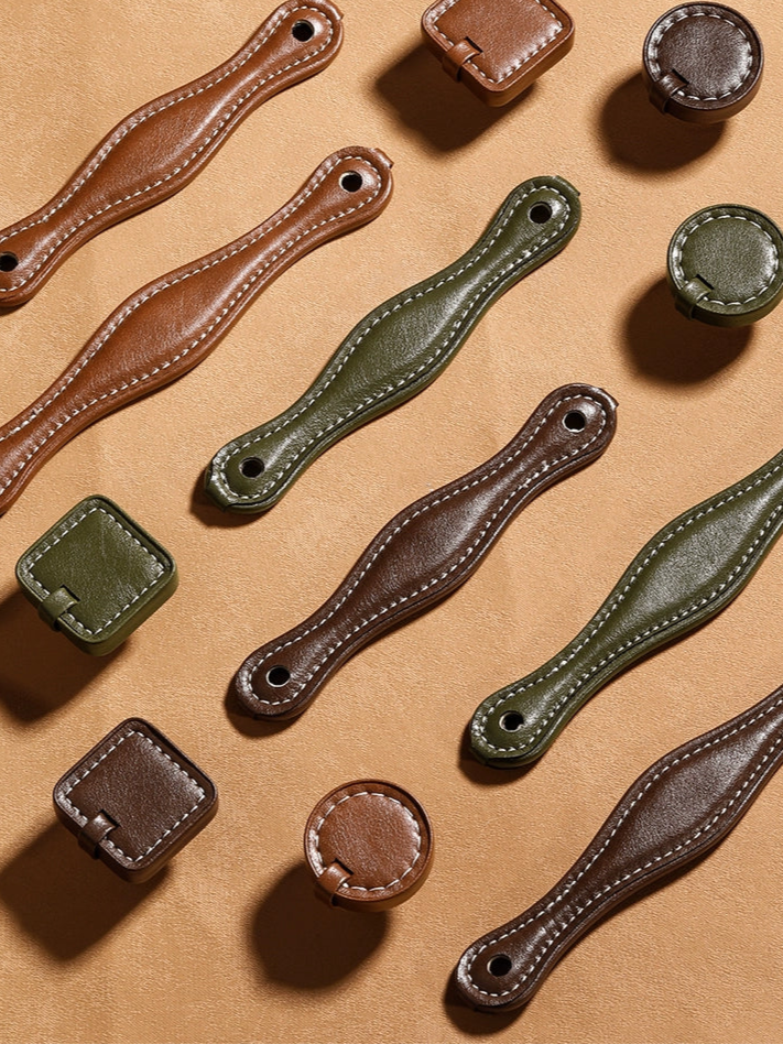 Brown and olive green leather-wrapped drawer pulls and cabinet handles in various shapes.  Stitched leather, metal mounting holes.  Modern farmhouse style.