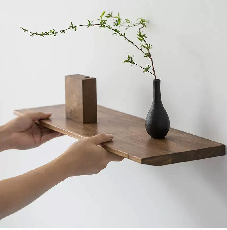 Hands installing a thick, solid wood floating shelf against a white wall, displaying a vase and block.
