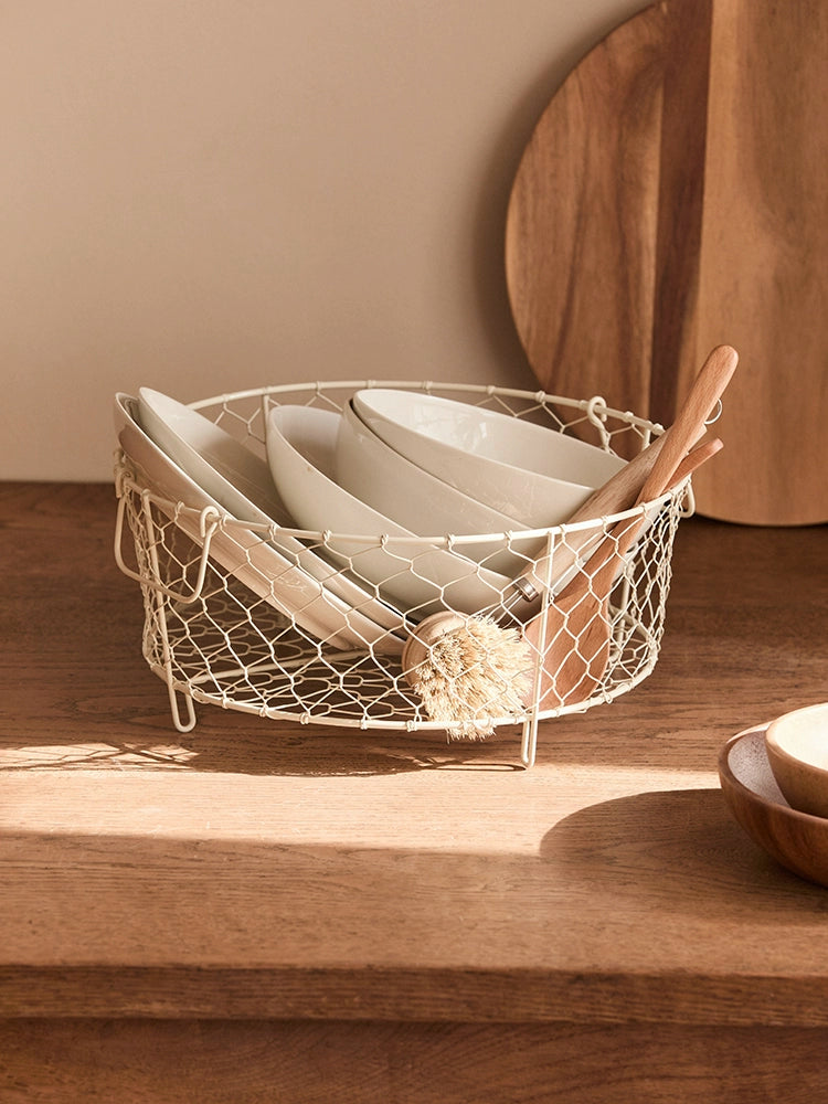 Beige wire dish basket with handles, holding white bowls and wooden utensils. Kitchen storage.
