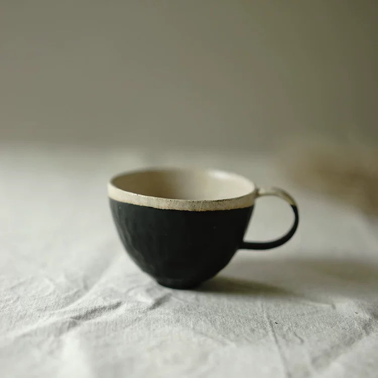 A rustic handcrafted ceramic cup with a matte black textured base and a natural cream-colored interior on a white cloth.