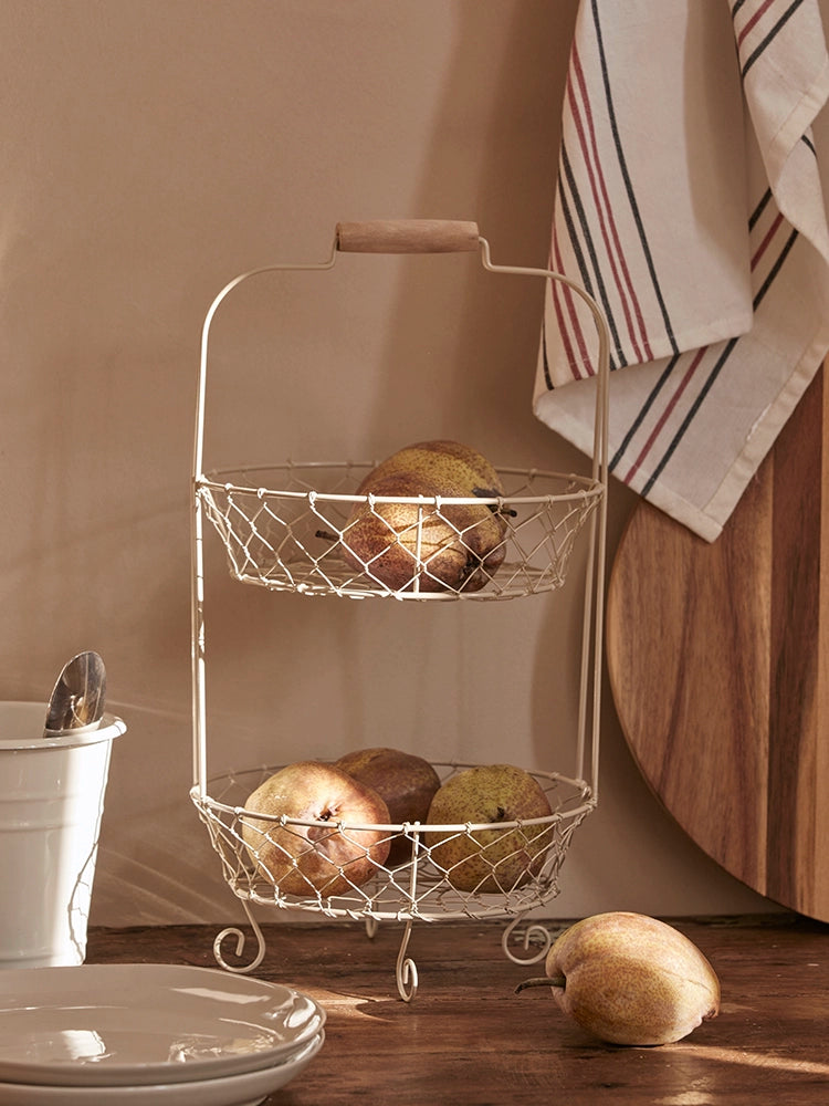 Two-tiered wire fruit basket stand, light beige wire, wooden handle, rustic kitchen decor.  Fruits in wire baskets.