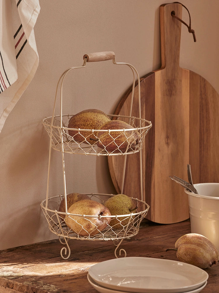 Two-tiered wire fruit basket stand, light beige wire, wooden handle, filled with pears. Rustic kitchen decor.