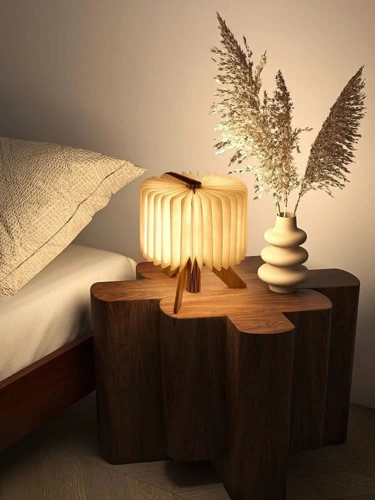 Warm light glows from folding portable paper lamp on a rustic wooden side table next to a bed.