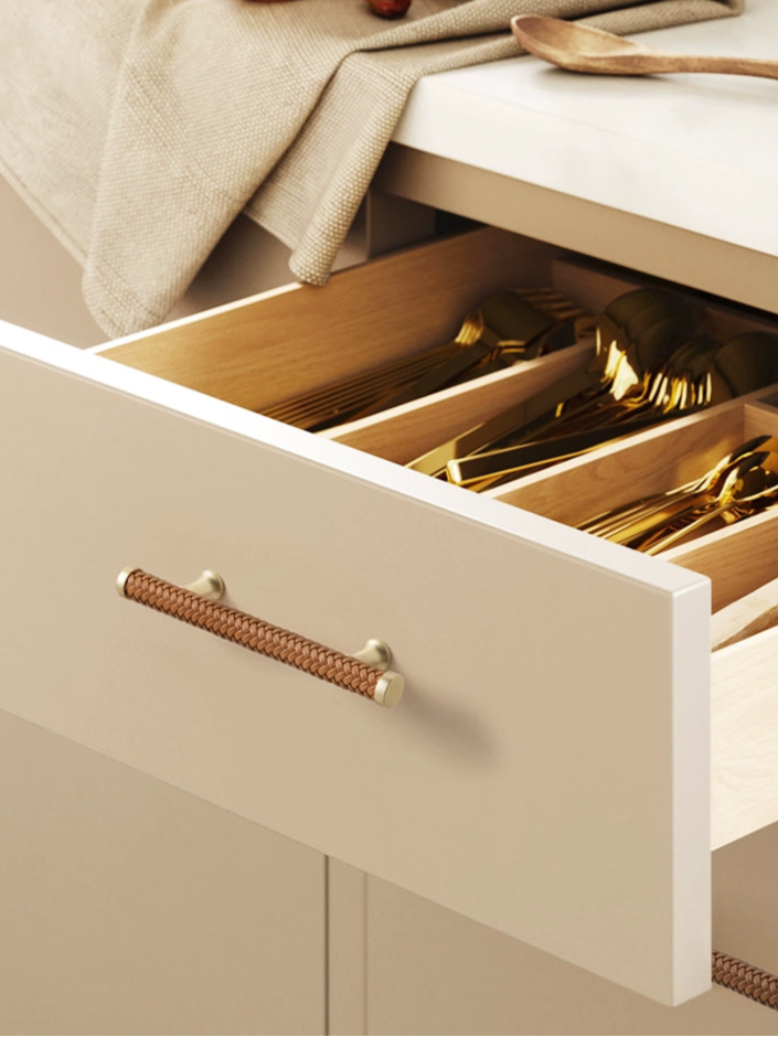 Woven leather drawer pulls in gold tone on a beige cabinet, showing golden cutlery inside.