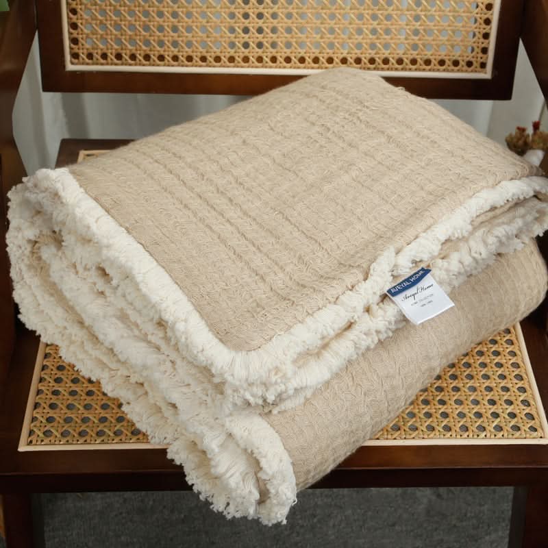 Beige linen cotton throw blanket with tassels, folded on a chair.