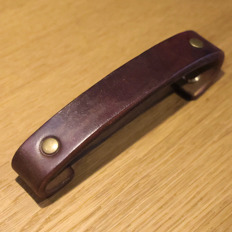 Rustic brown leather drawer pull with brass studs on a wooden surface.