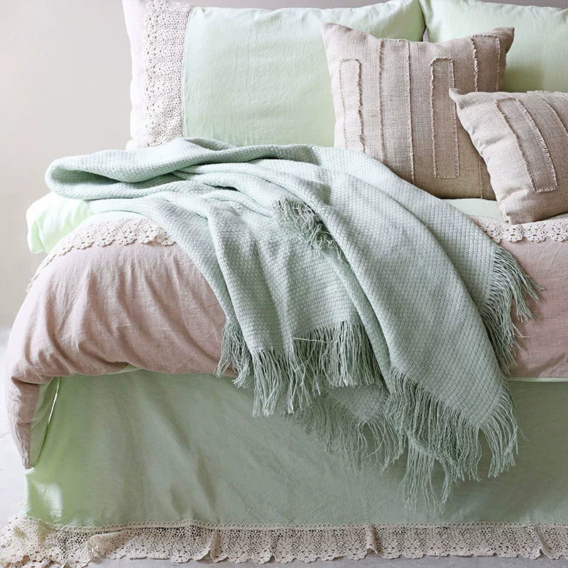 Light green linen throw blanket with fringes draped on a bed.