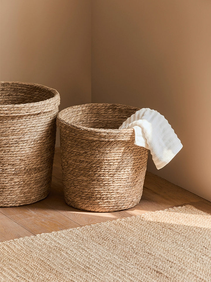 Two natural rattan storage baskets, round and square, on a light brown wooden floor.  Beige colored baskets with woven texture.  A white towel is inside the round basket.