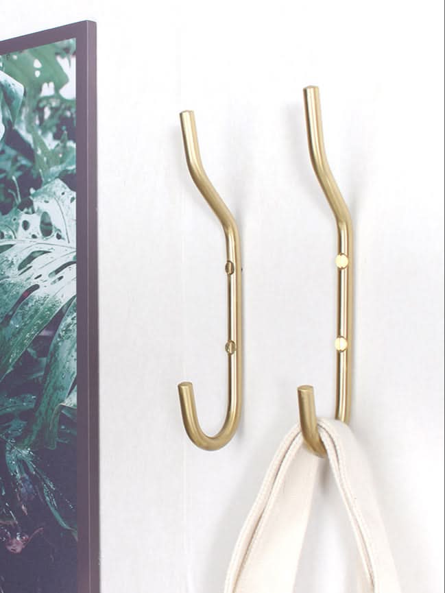 Minimalist brass wall hooks mounted on white wall with green art to the left.