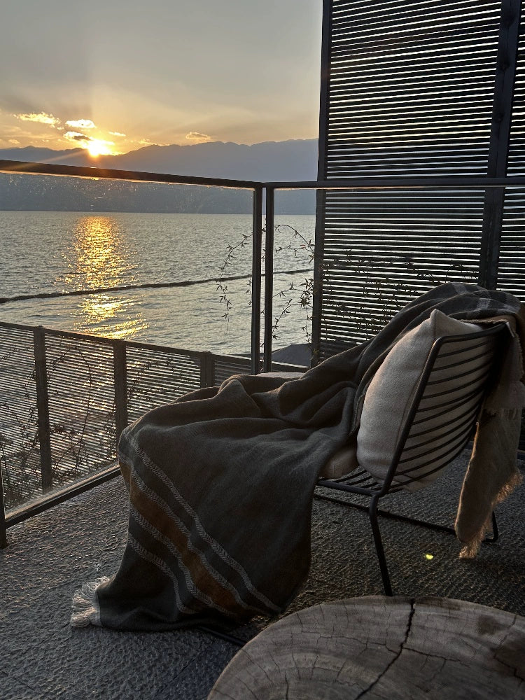 Cozy striped linen throw blanket in multicolor draped on a chair, with a scenic sunset over water.